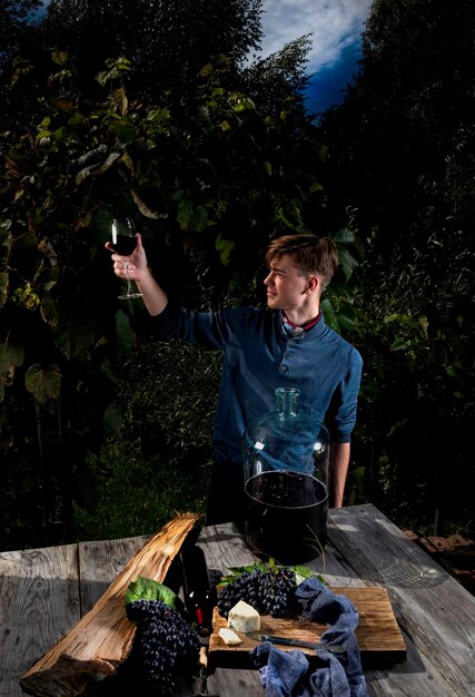 A young man testes homemade red wine from a glass against the background of a vineyard