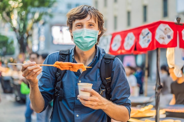 Young man tourist in medical mask eating typical korean street food on a walking street of seoul