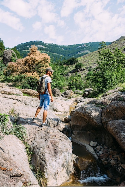 Young man traveler walking alone by a mountain river with rocky summer landscape