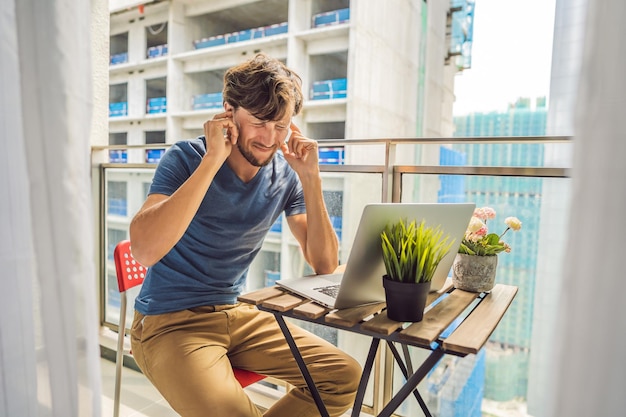 Young man trying to work on the balcony annoyed by the building works outside Noise concept Air pollution from building dust