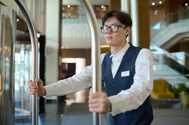 Young man in uniform of bellboy pushing cart with luggage along lounge