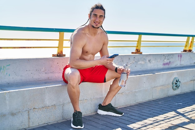 Young man using smartphone sitting on bench at seaside