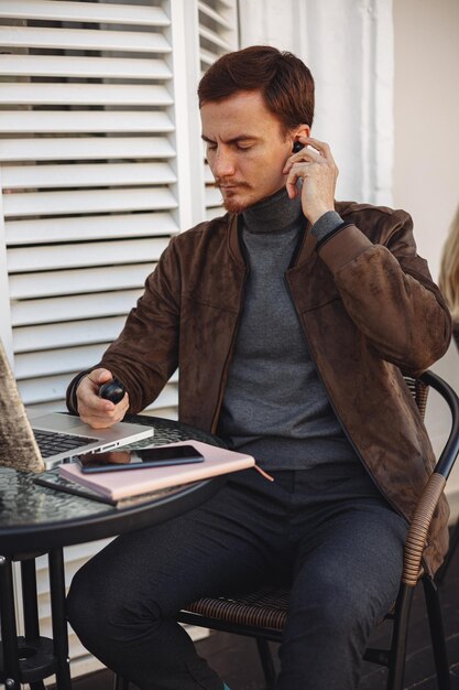 Young man using wireless earphones and preparing for video call via laptop in city cafe 