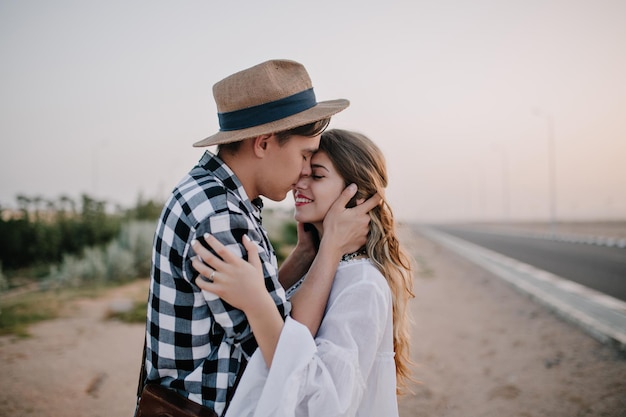 Young man in vintage hat gently holding girlfriend's face and going to kiss her standing near the road at the dawn. Beautiful couple in love cute hugging outside enjoys romantic evening date.