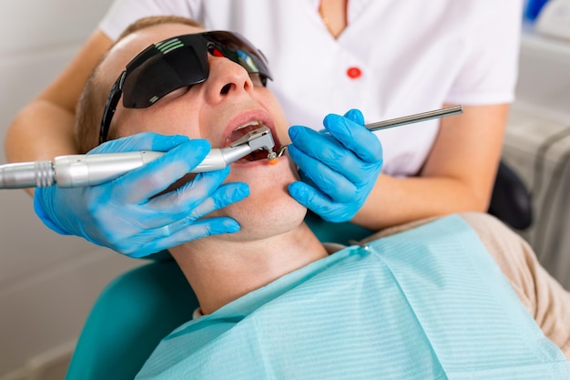 Young man visiting the dental office Dentist hands in protective gloves using ultrasound to clean patient's teeth Closeup of open mouth