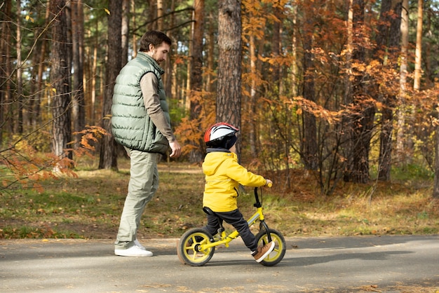 Young man walking by his little son in safety helmet and casualwear riding balance bicycle along wide road in park while enjoying chill at leisure