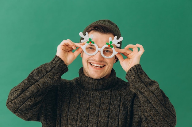 A young man in a warm sweater a cap and carnival glasses isolated on a green background