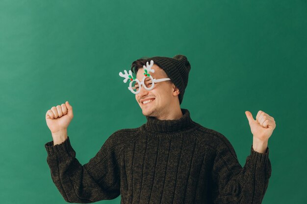 A young man in a warm sweater a cap and carnival glasses isolated on a green background