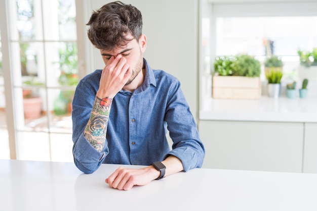 Young man wearing casual shirt sitting on white table tired rubbing nose and eyes feeling fatigue and headache Stress and frustration concept