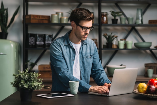 Young man wearing eyeglasses using laptop on kitchen counter