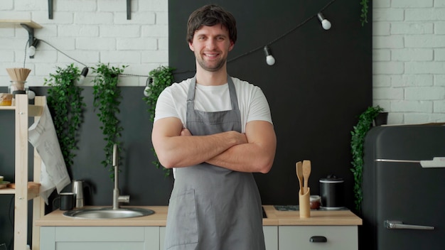 Young man wearing gray apron smiling and crossing arms standing in her kitchen