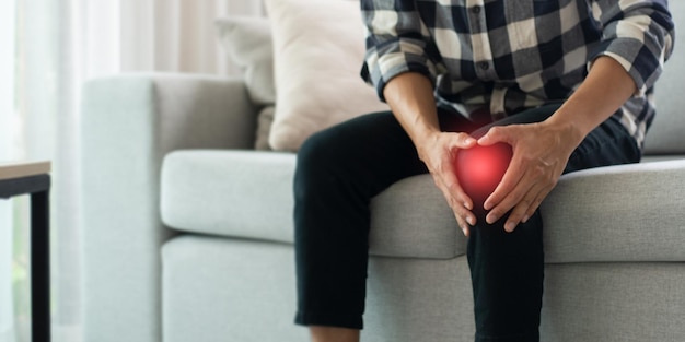 A young man wearing a plaid shirt and black pants sits on the sofa in the living room at home He felt pain in his knee area so he used both hands to hold his knee