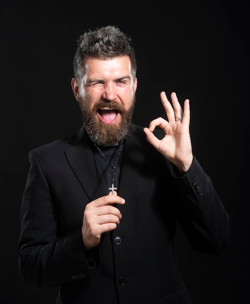 Young man wearing priest uniform standing over white background doing ok gesture with hand smiling, eye looking through fingers with happy face.