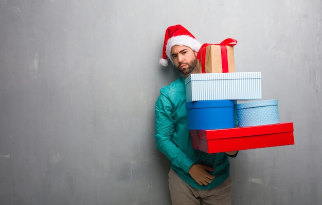 Photo young man wearing a santa hat holding gifts looking straight ahead