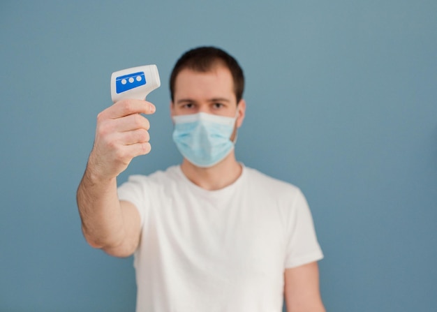 Young man wearing surgical mask is using infrared thermometer on a blue background