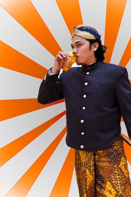 Photo young man wearing traditional clothing while drinking fresh jamu against pattern wall