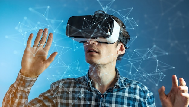 A young man wearing virtual reality glasses watching at a 3D visualization in an abstract polygonal grid on a blue background in a futuristic style