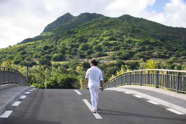 A young man in white clothes walks along the road against the background of a mountain