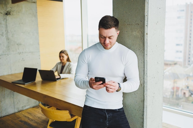 A young man in a white pullover is standing in a spacious office and talking on the phone A man talks to colleagues via video link