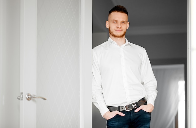 A young man in a white shirt and jeans stands in the doorway and looks into the camera