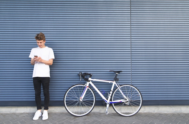 Photo young man in a white shirt standing near the road bike and uses the smartphone