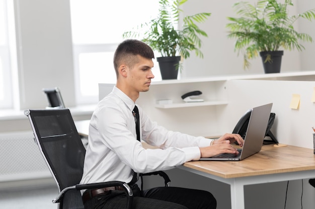 Young man in a white shirt with a black tie at the desk at the bright office working on a laptop