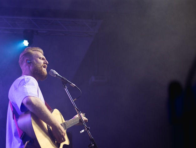 A young man in a white T-shirt with a beard holds an acoustic guitar in his hand and sings into a microphone stands on the stage