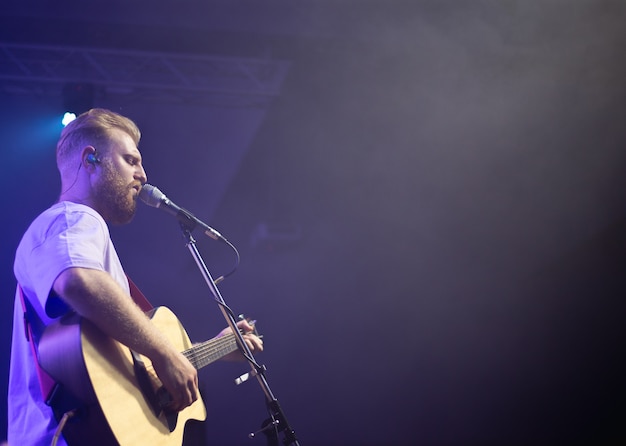 A young man in a white T-shirt with a beard holds an acoustic guitar in his hand and sings into a microphone stands on the stage