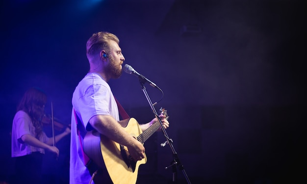 A young man in a white T-shirt with a beard holds an acoustic guitar in his hand and sings into a microphone stands on the stage