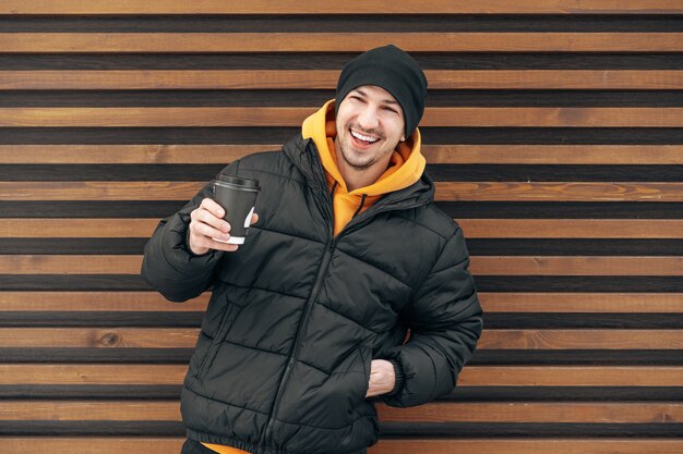 Photo young man in winter clothes standing with coffee to go in a street against wooden wall