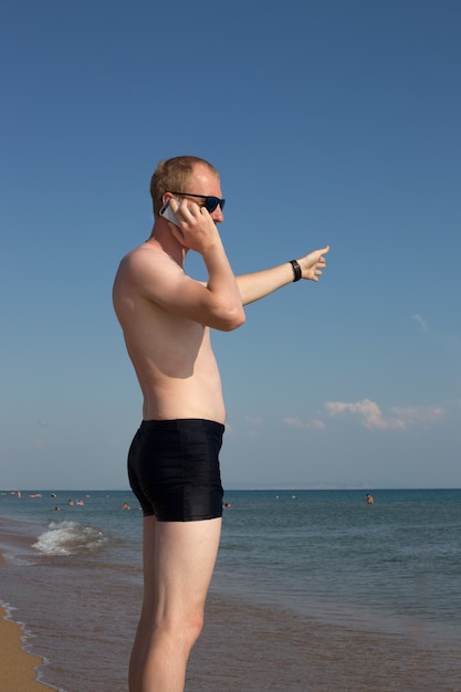 A young man with a bare torso is standing on the seashore and talking on the phone