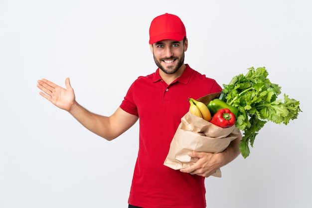 Young man with beard holding a bag full of vegetables isolated on white extending hands to the side for inviting to come