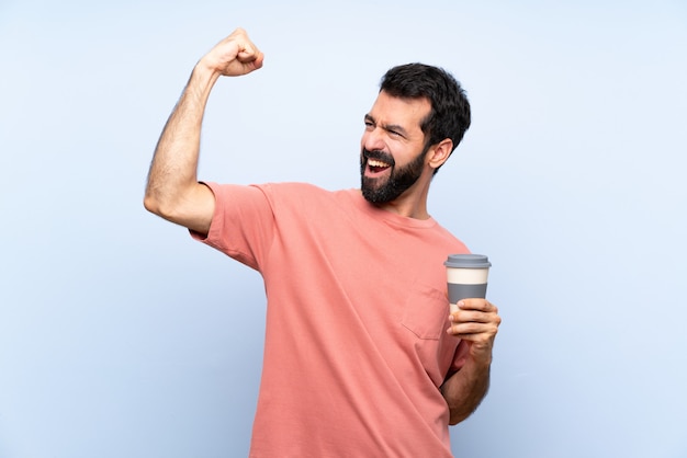 Young man with beard holding a take away coffee over isolated blue   celebrating a victory