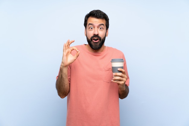 Photo young man with beard holding a take away coffee over isolated blue   surprised and showing ok sign