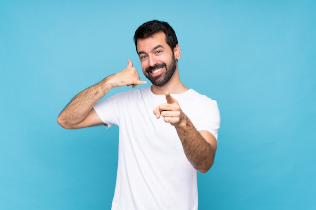 Young man with beard  over isolated blue background making phone gesture and pointing front