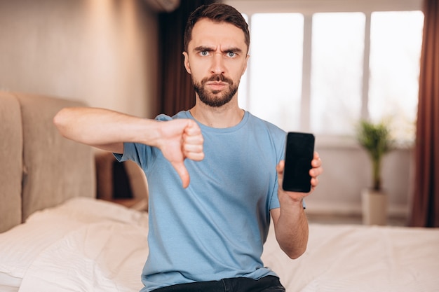 Young man with beard sitting in bed and shows disappoint and does not like using phone after sleep while