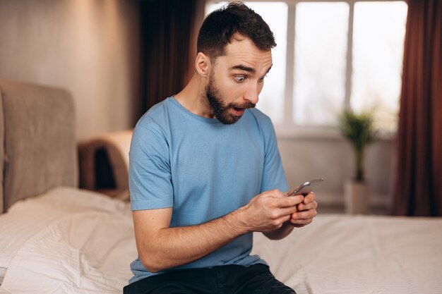 Young man with beard surprised shocked whilst received sending message on phone while sitting in bed