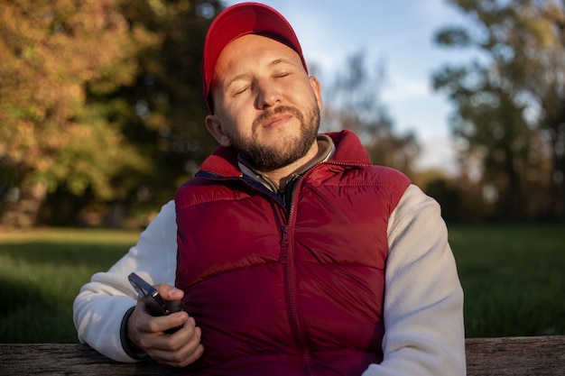 Young man with a beard with a phone sitting on a park bench