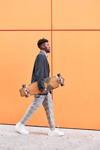 young man with blazer and skateboard walking down the street against an orange background