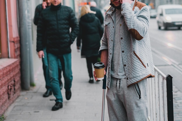 Young man with cane in casual clothing stands on the city's street Stick Crowd Rush hour Wait Cup Hold Coffee Life Leg Boy Step Walk Guy Disease Man Alone Urban