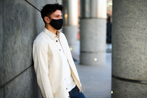 Young man with covid or coronavirus mask leaning against a wall in a city