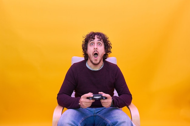 Young man with curly hair playing video games over isolated yellow background. Surprised looking to the camera.