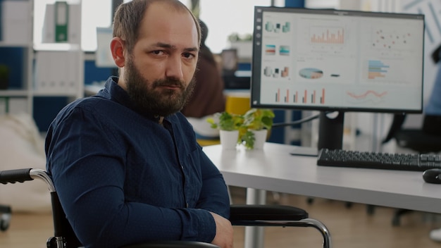 Young man with disabilities, invalid, handicapped disabled paralized immobilized looking at camera upset, tired sitting in wheelchair in business office room, working at financial project with team.