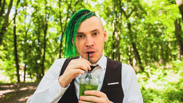 Young man with dreadlocks drinking refreshing lemonade in forest park Stylish hipster in business suit with green hairstyle drinking soft drink through straw quenching thirst in summertime