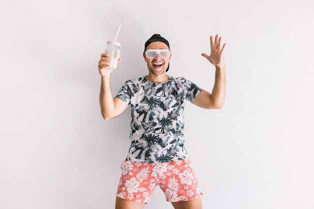 Young man with a floral shirt, cap and glasses in summer drinking a cocktail with a straw and making joyful gestures, in daylight on a white wall