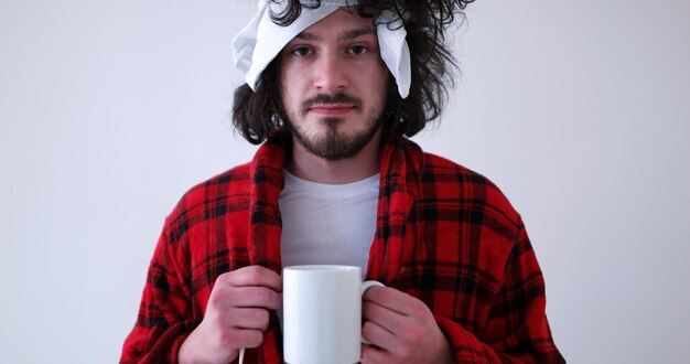 young Man with flu and fever wrapped holding cup of healing tea isolated over white