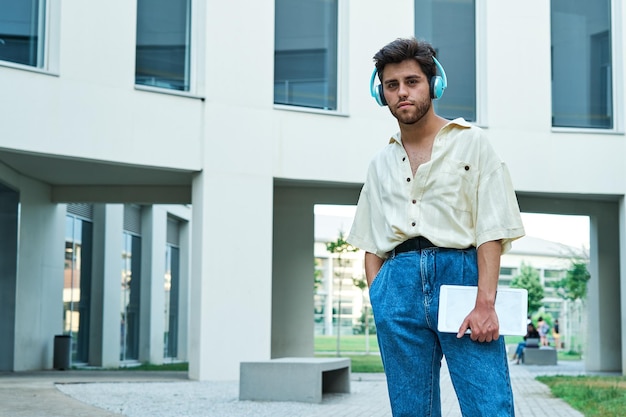 Young man with headphones holding digital tablet standing and looking at camera outside building