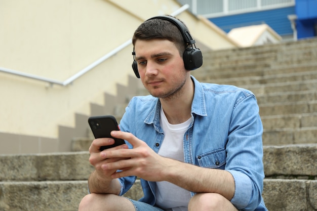 Young man with headphones sits on the stairs and looks at his phone