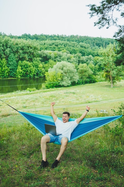 A young man with a laptop sits in a hammock in nature and works remotely Work in nature during vacation