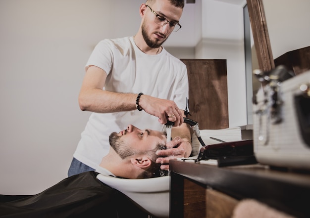 Young man with trendy haircut at barber shop. Barber washes customer head.